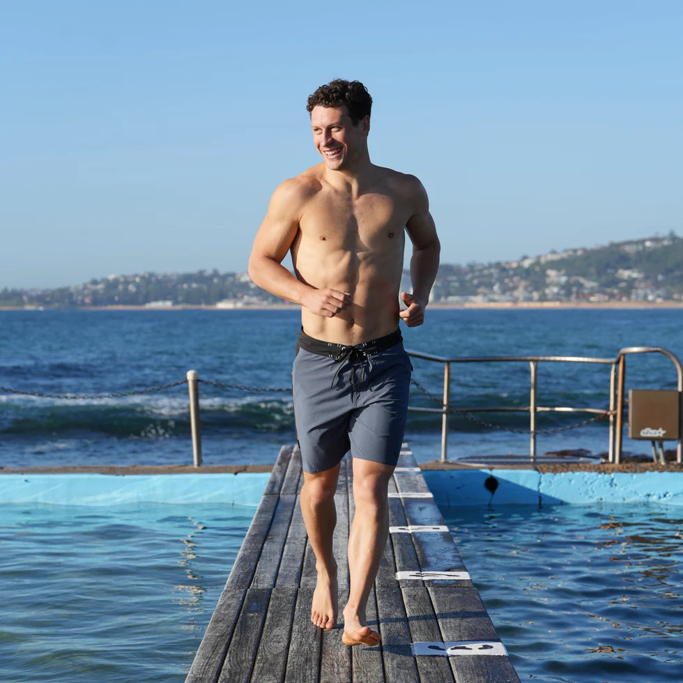 Vaikobi Newport Boardie (VK-262) Man walking on a wooden platform by the ocean with a clear blue sky.